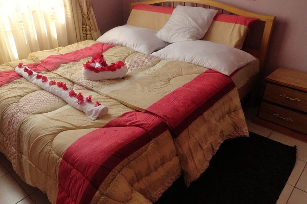 A neatly made bed with a beige and red bedspread, white pillows, and a red floral decoration.