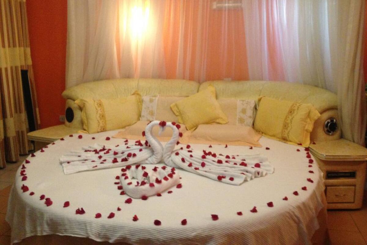 A bed with a swan-shaped towel arrangement and rose petals, a yellow sofa, and a nightstand with a lamp.