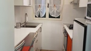 Eine kleine Küche mit weißen Schränken, einem Spülbecken, einem Herd und einem Fenster mit transparenten Vorhängen.