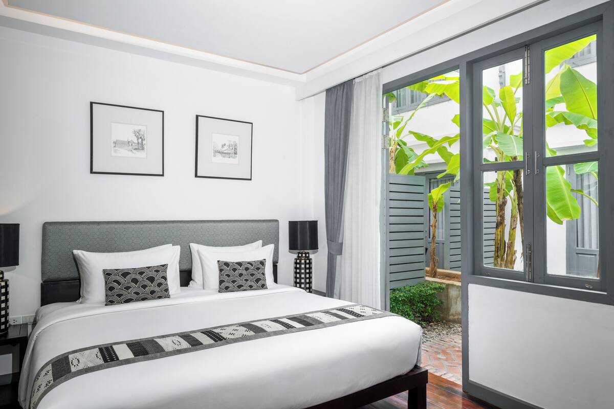 A neatly arranged bedroom with a large bed, two framed pictures on the wall, and a view of greenery through a window.