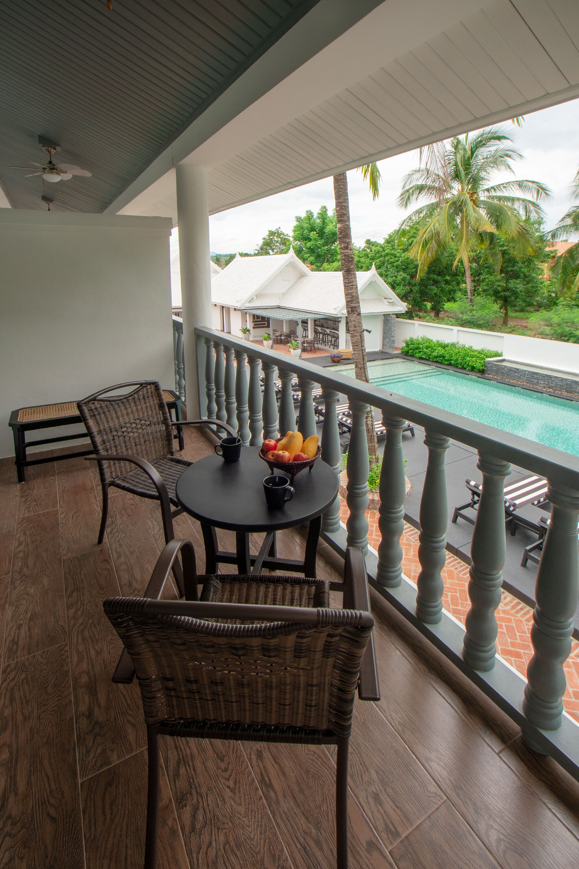 A balcony with wicker furniture, a table with fruit, and a view of a swimming pool and palm trees.