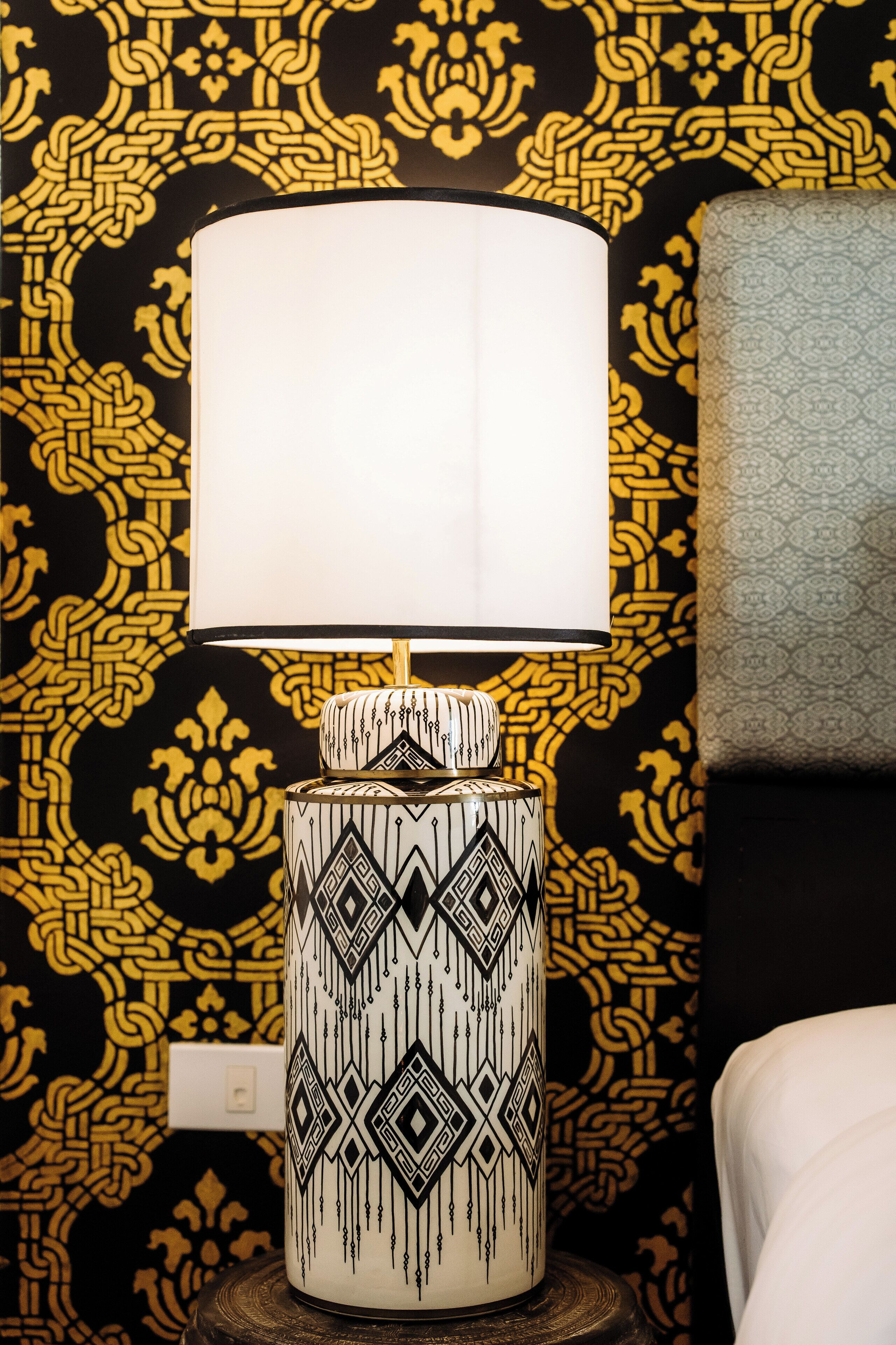 A patterned lamp with a white shade on a wooden table against a black and gold patterned wall.