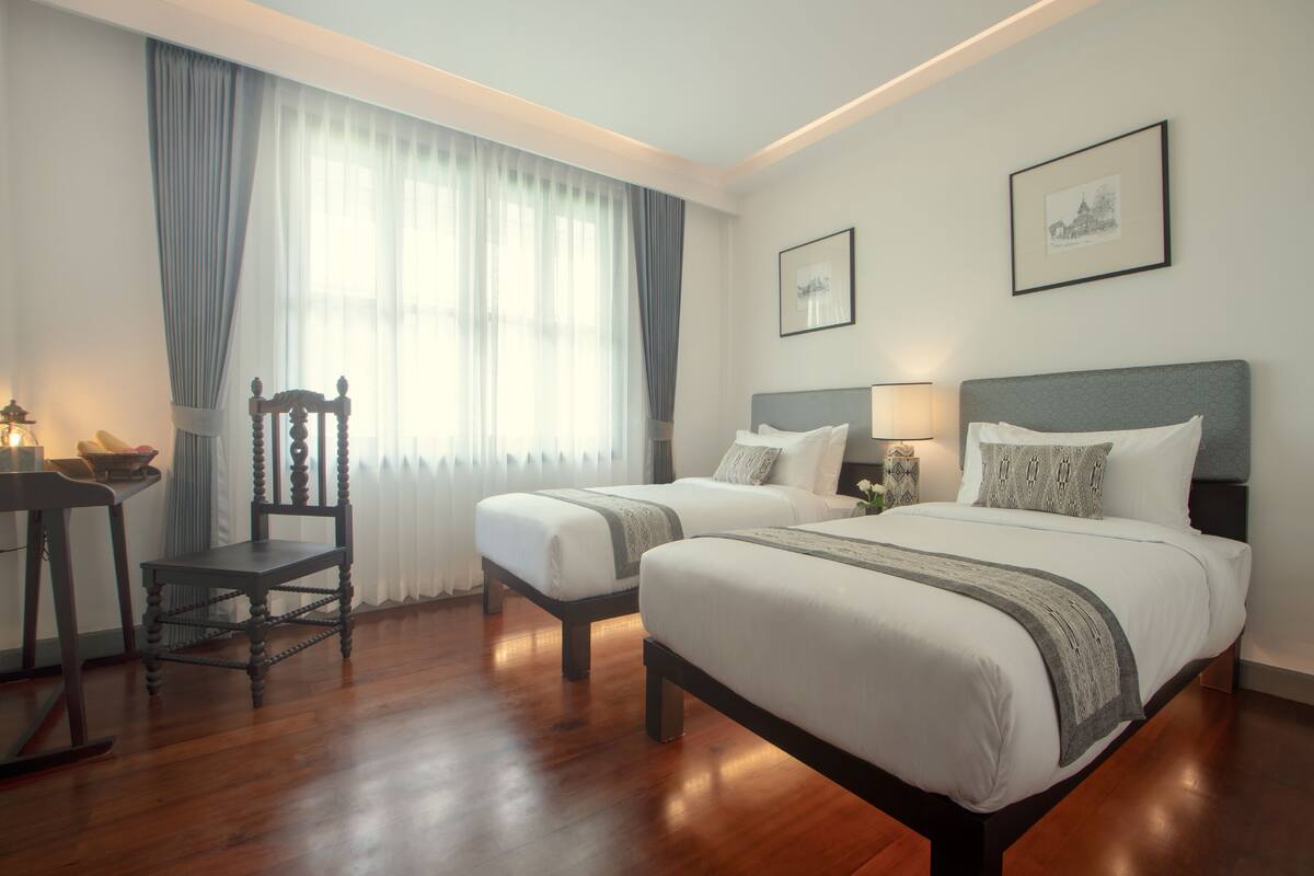A hotel room with two beds, a wooden chair, a table with a lamp, and a window with curtains.