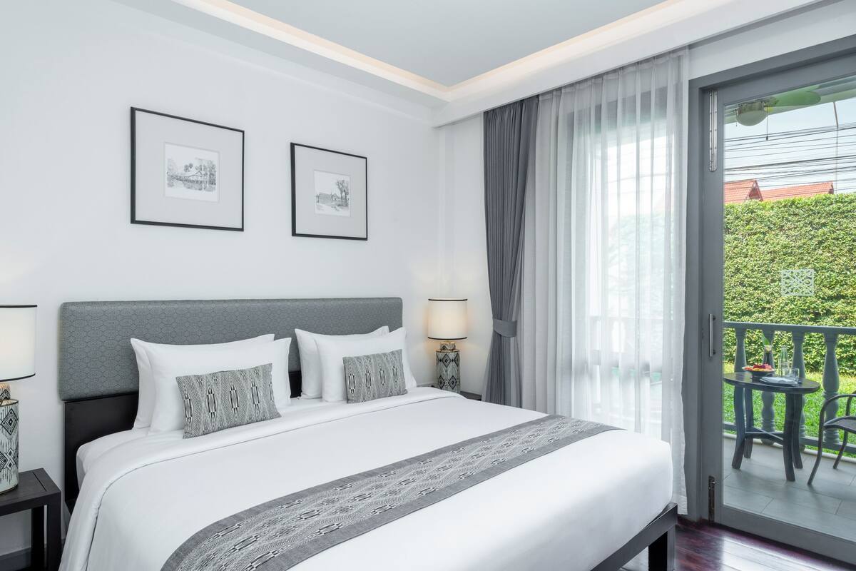 A modern hotel room with a large bed, two framed pictures on the wall, a balcony with a table and chairs, and a view of greenery outside.