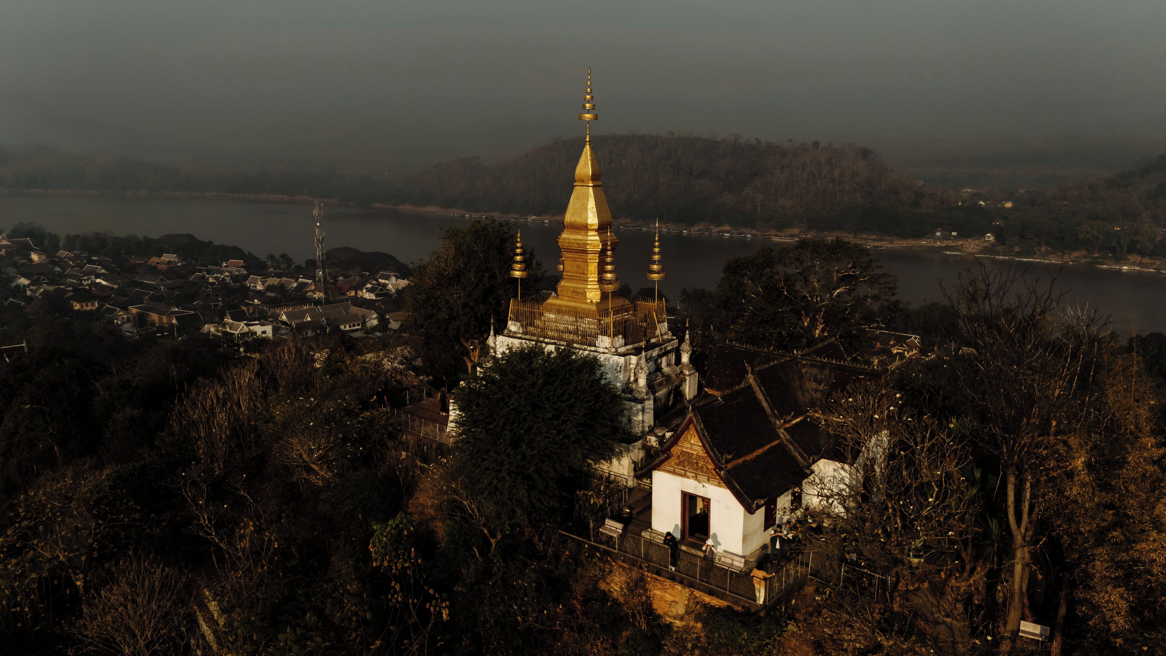 A temple complex with a prominent golden spire, surrounded by dense forest and overlooking a body of water.