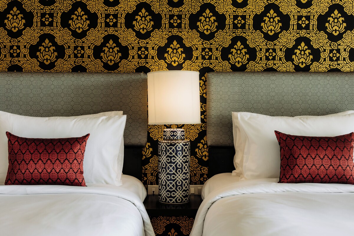 A hotel room with two beds, a nightstand with a lamp, and a patterned wallpaper.