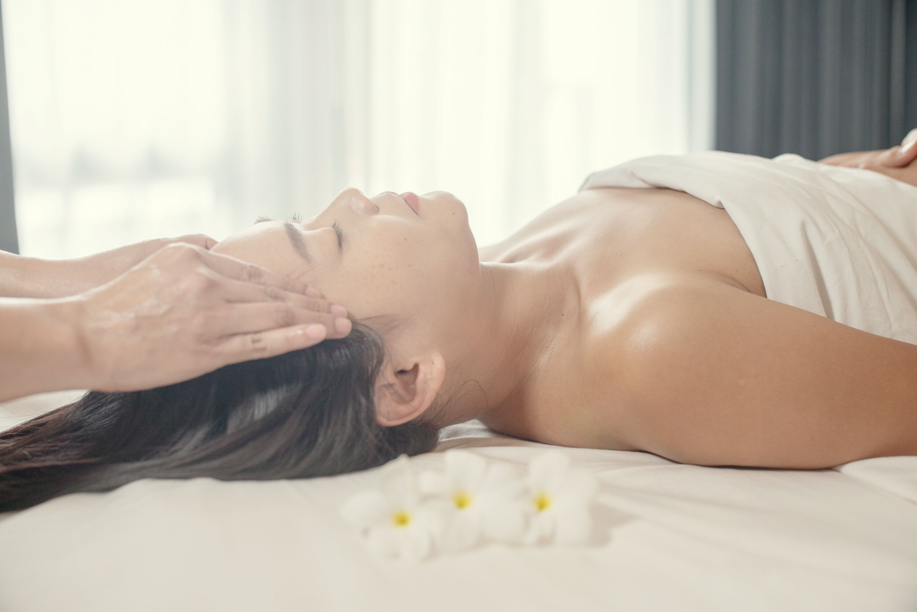 A person lying on a bed with white flowers, receiving a facial treatment.