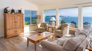 A spacious living room with a large window overlooking the sea, a wooden cabinet, and a beige sofa. - Orcas Island, Mt. Baker Beach House, #289 (Eastsound)