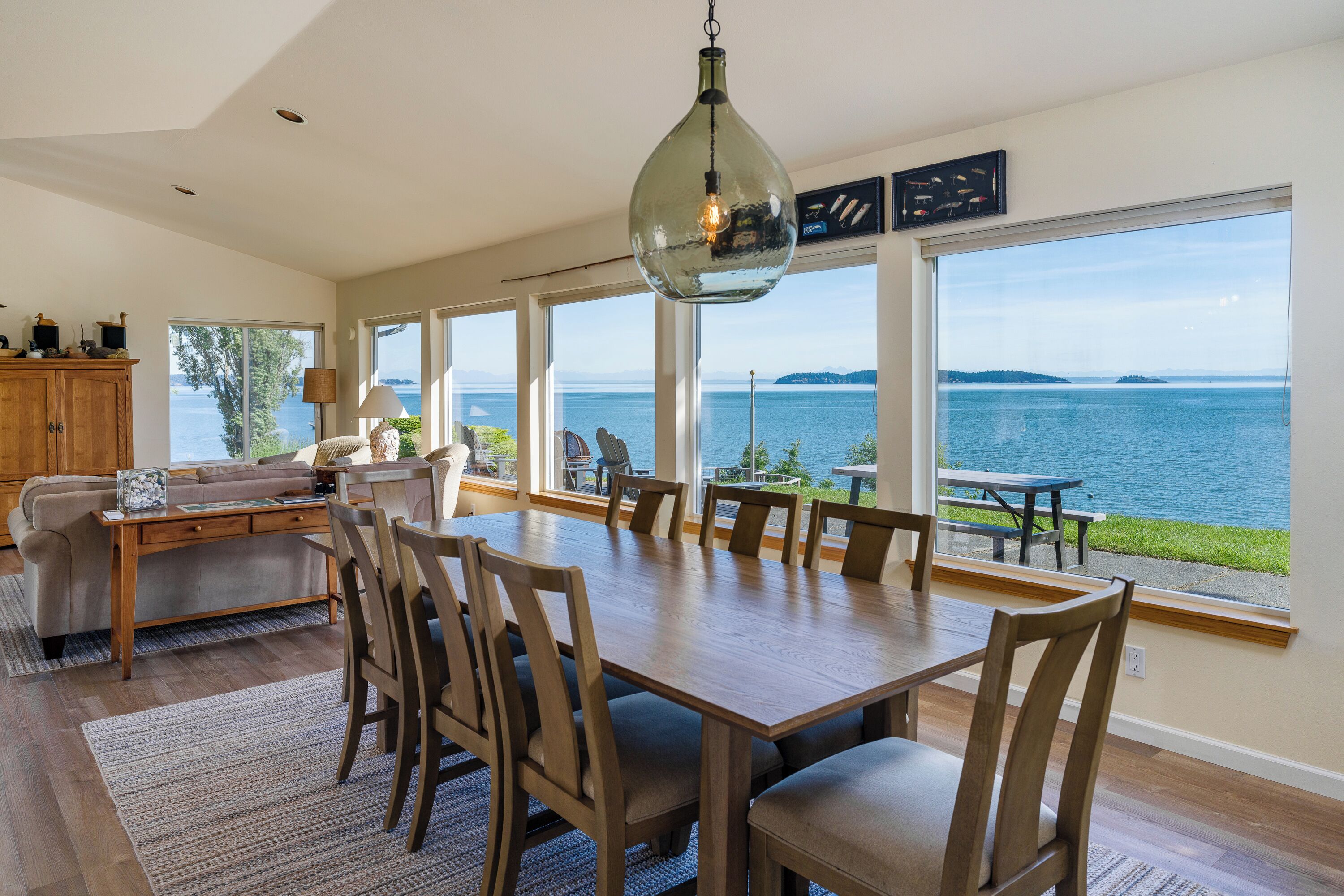 A spacious dining area with a large table and chairs, a hanging light fixture, and large windows offering a view of the water.