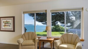 A room with two armchairs, a small table, a lamp, a framed picture of fish, and large windows with a view of the sea and trees. - Orcas Island, Mt. Baker Beach House, #289 (Eastsound)