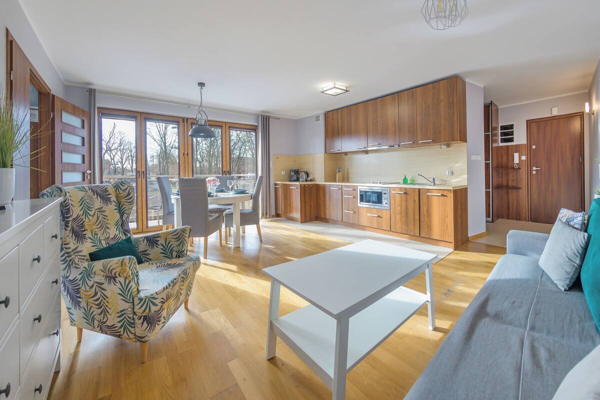 A modern living room with a sofa, a dining area with chairs, a kitchen with wooden cabinets, and a TV on a cabinet.