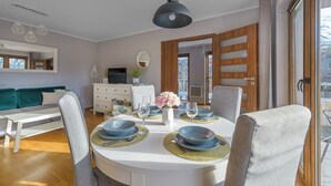 Comedor con mesa redonda dispuesta para cuatro personas, rodeada de sillas. Hay un televisor sobre un mueble blanco, un sofá al fondo y una puerta que da al exterior.