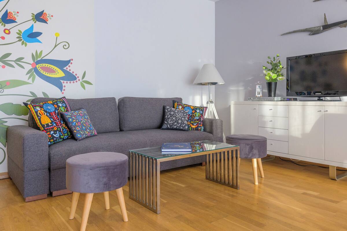 A modern living room with a grey sofa, colorful pillows, a glass coffee table, and a white cabinet with a flat-screen TV.