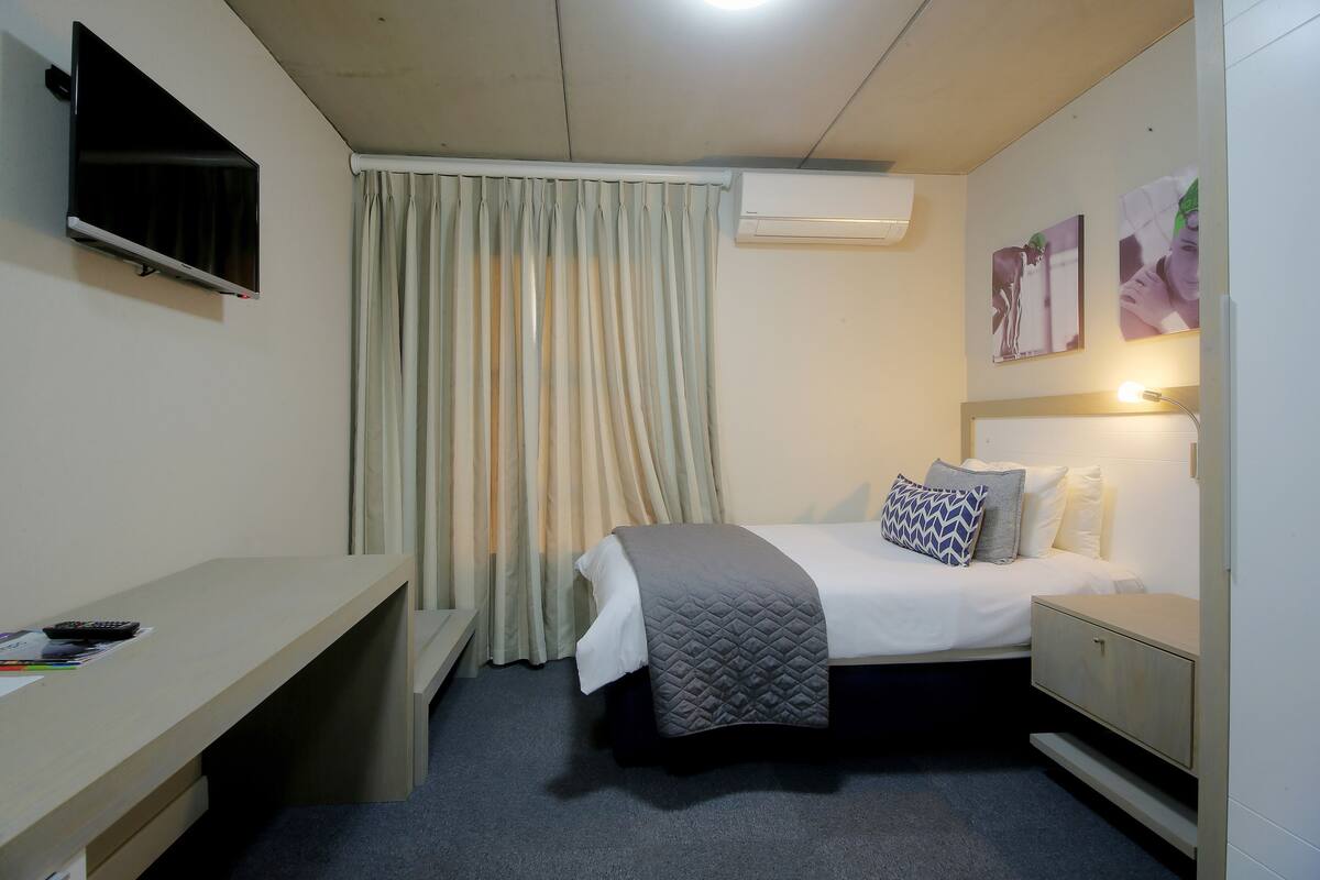 A hotel room with a bed, bedside table, TV, air conditioning unit, and a curtain.