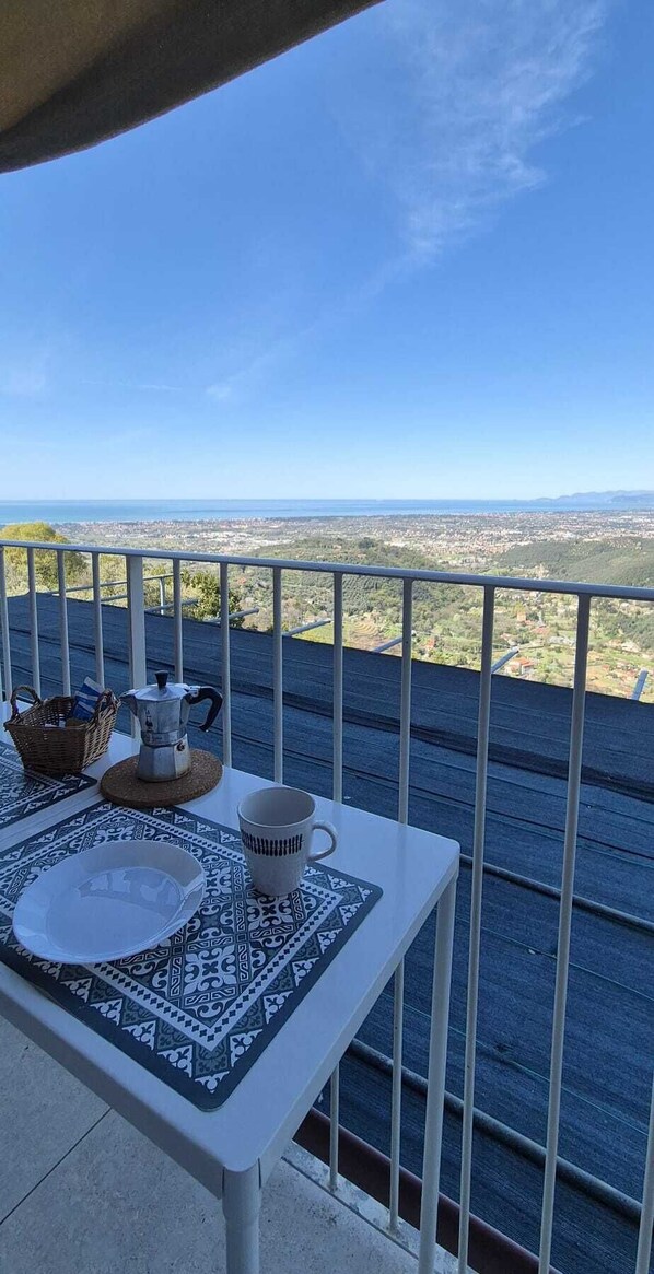 A balcony with a table set for two, a coffee pot, and a cup, overlooking a cityscape.