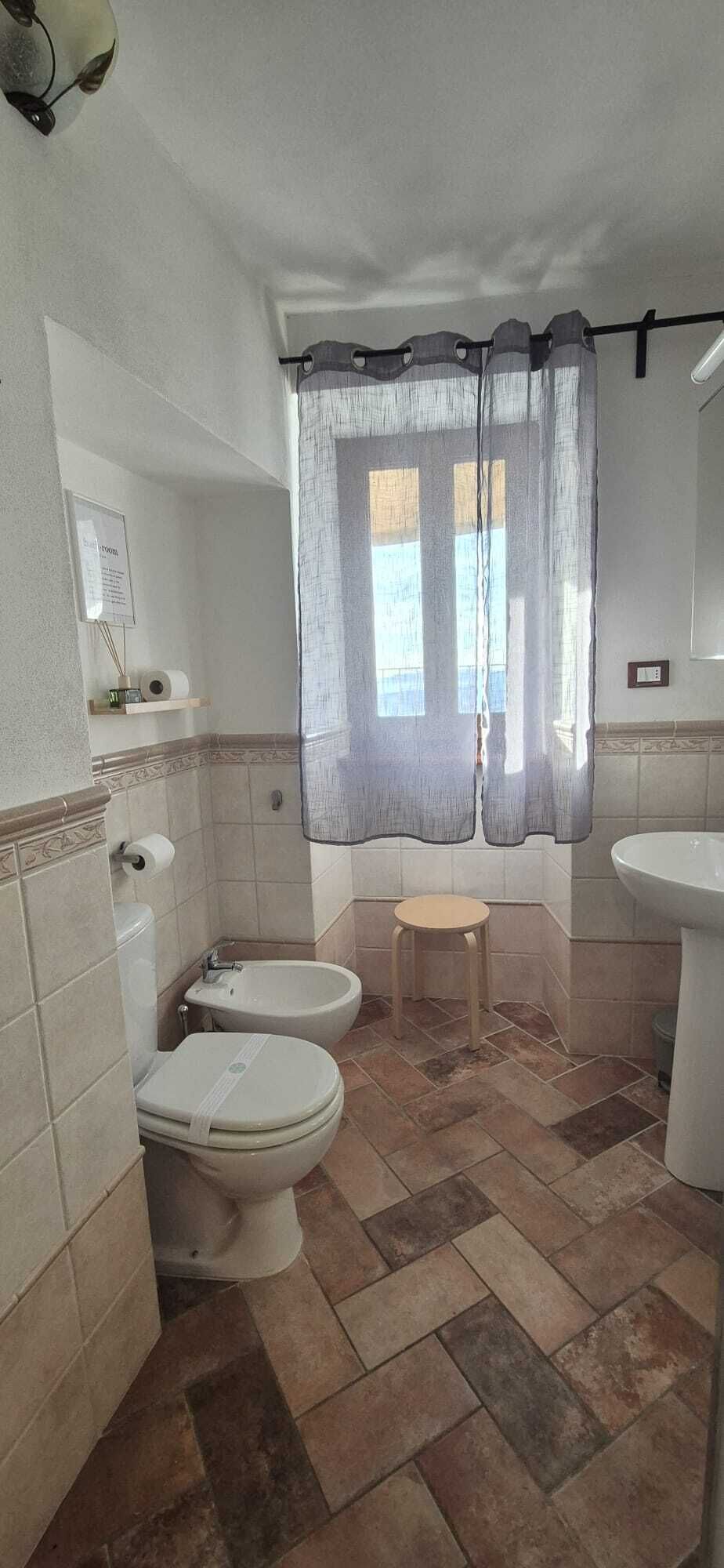 Ein Badezimmer mit zwei Toiletten, einem Bidet, einem Waschbecken und einem Fenster mit Vorhang.