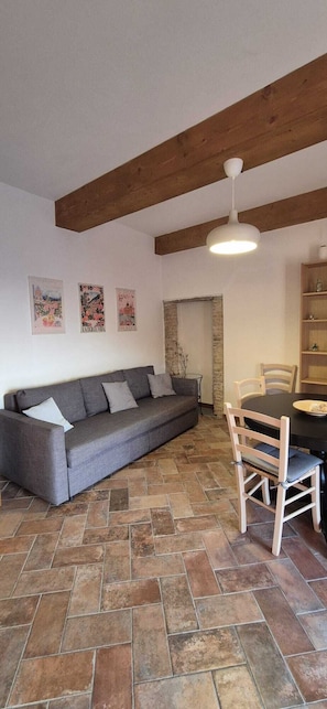 A room with a grey sofa, a dining table with chairs, a hanging light fixture, and a stone floor.