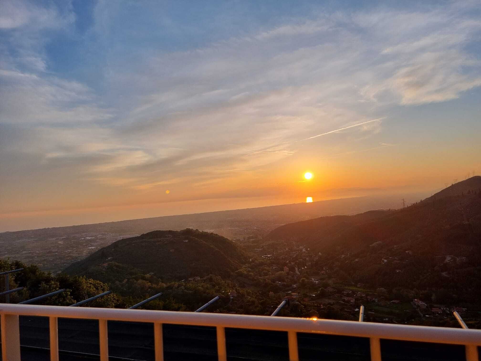 Zonsondergang vanaf een balkon met een hek, met uitzicht op een vallei en verafgelegen heuvels.