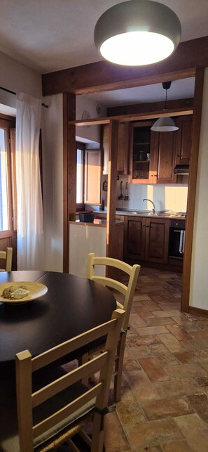A dining area with a table and chairs, a kitchen with wooden cabinets, a hanging light fixture, and a tiled floor.
