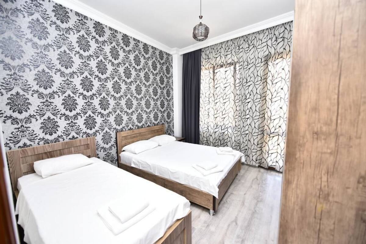 A hotel room with two beds, patterned wallpaper, a wooden headboard, and a large window with curtains.