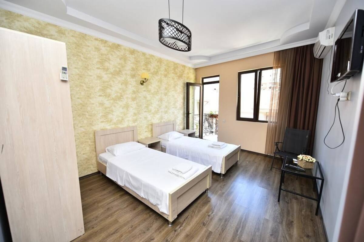 A hotel room with two single beds, a wooden wardrobe, a small table with a vase, a TV, and a window with curtains.