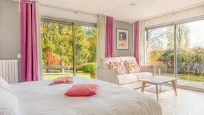 A bedroom with a large bed, a sofa, and a table. There is a view of a garden through the windows. - La Maison de L'orbière (Forcé)