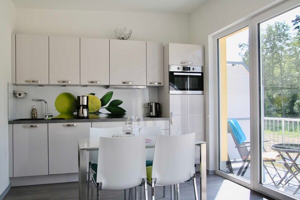 A modern kitchen with white cabinets, a dining table with chairs, and a view of the outdoors through large windows.