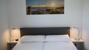 A bedroom with a bed, two bedside lamps, a nightstand, a wall-mounted picture of a sunset, and a power outlet. - *öddeker, Lidia und Dieter GM 69137 - Holiday apartment in the coastal forest - Baltic Sea SNR: (Gelbensande)