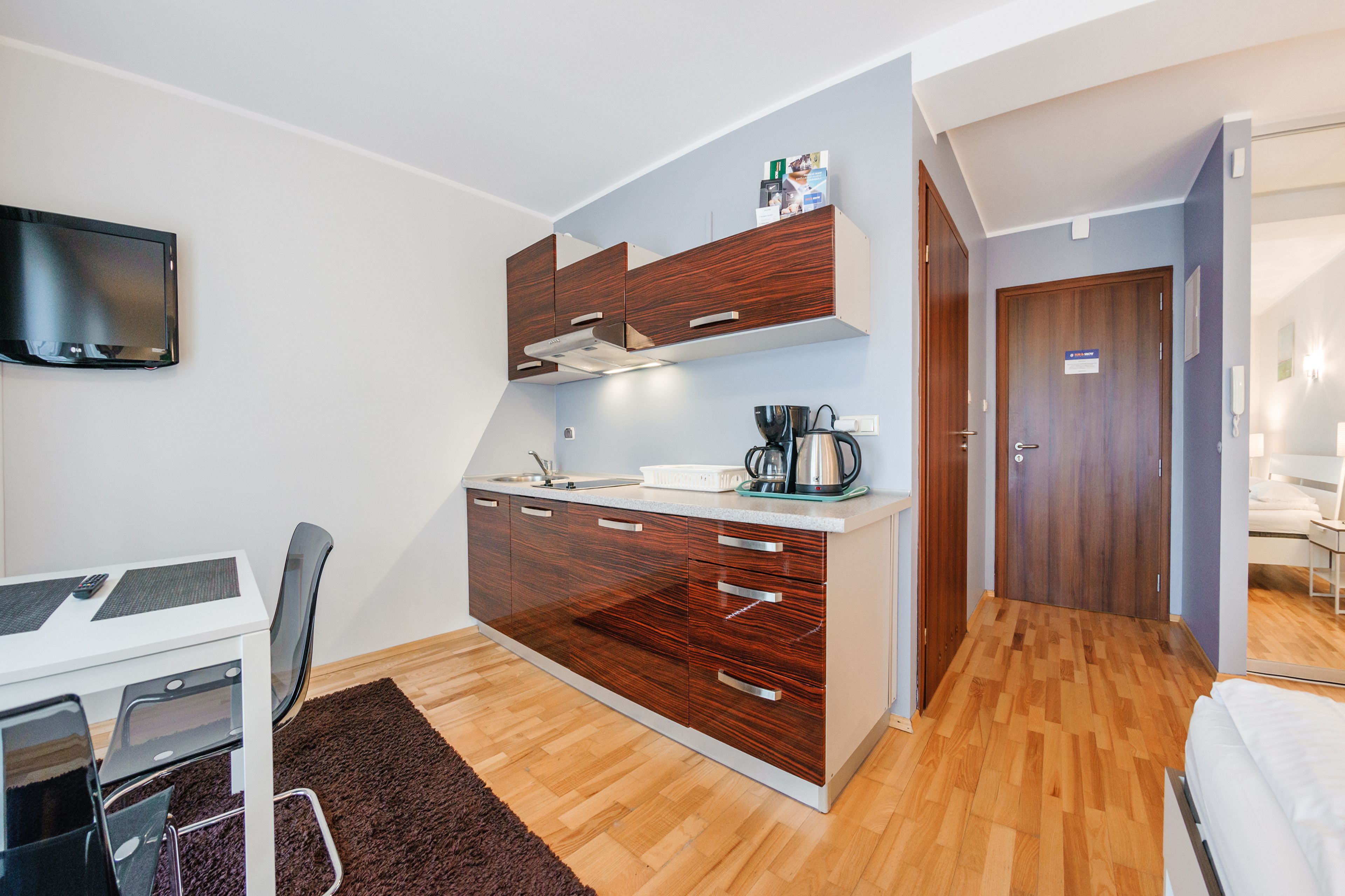 A compact kitchen with a built-in stove, a sink, and a coffee maker. There is a dining table with chairs and a TV mounted on the wall.