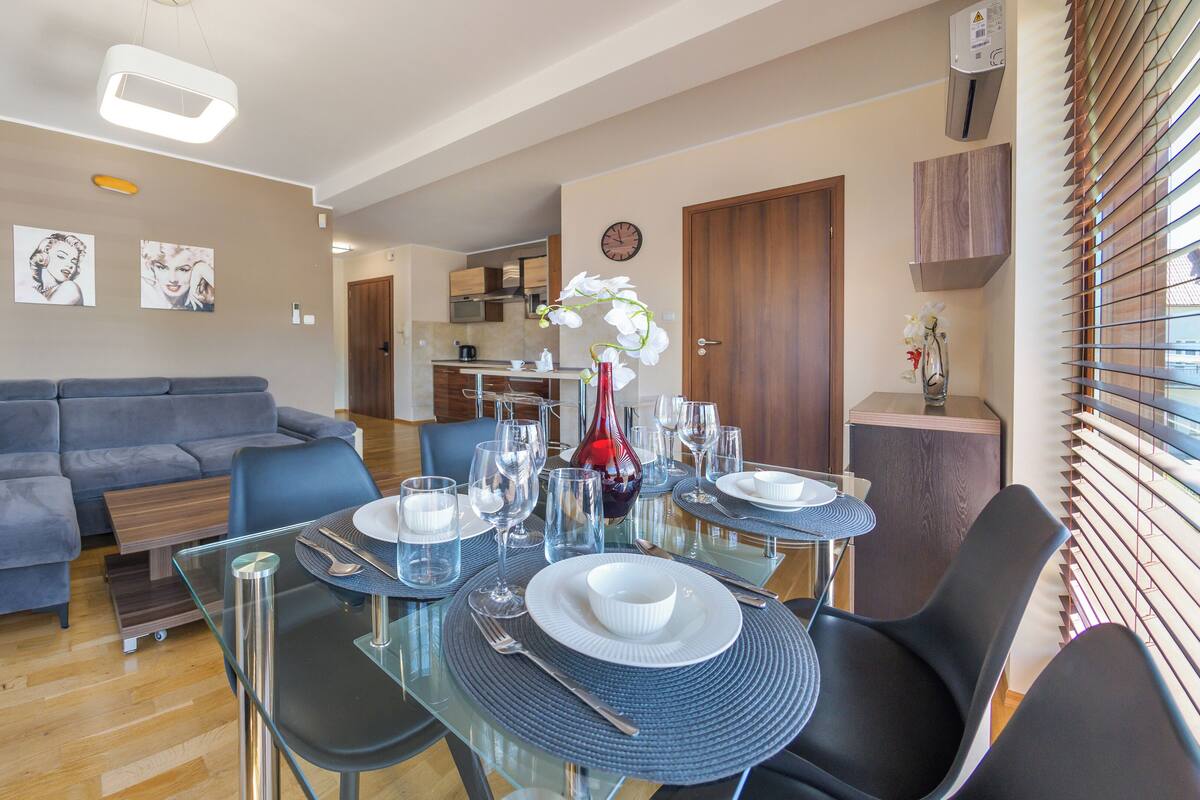 A modern living room with a grey sofa, a dining table set with plates, glasses, and cutlery, and a kitchen in the background.