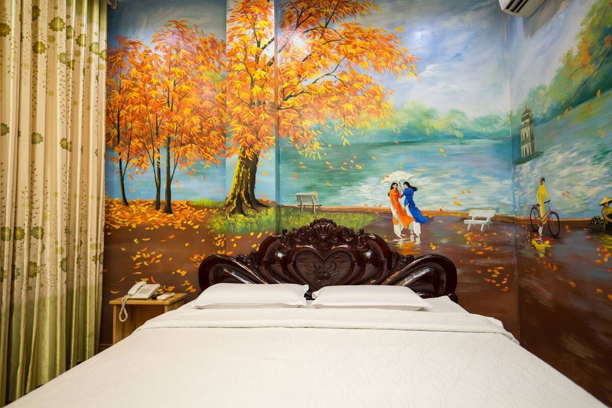 Une chambre à coucher comprenant un lit, une table de chevet, un téléphone et une fresque murale représentant un paysage lacustre avec des arbres et des personnes.