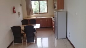 A compact kitchen with a dining area, a refrigerator, and a window with curtains. - Kami Executive Apartments (Kigali)