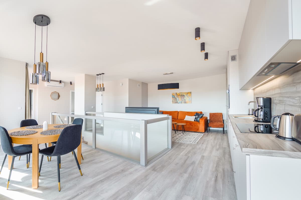 A modern kitchen with a central island, a dining area with a wooden table and chairs, and a living area with a sofa and a coffee machine.