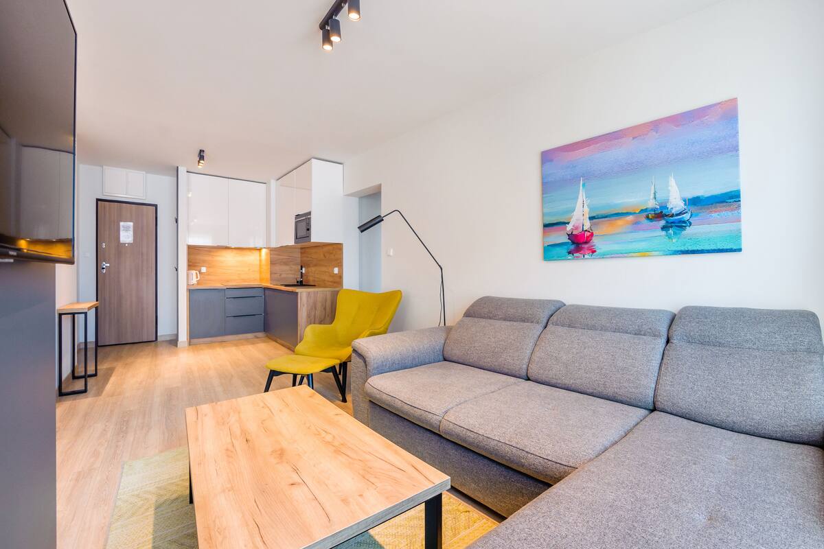 A modern living room with a grey sofa, a wooden coffee table, and a yellow chair. A painting of sailboats is on the wall.