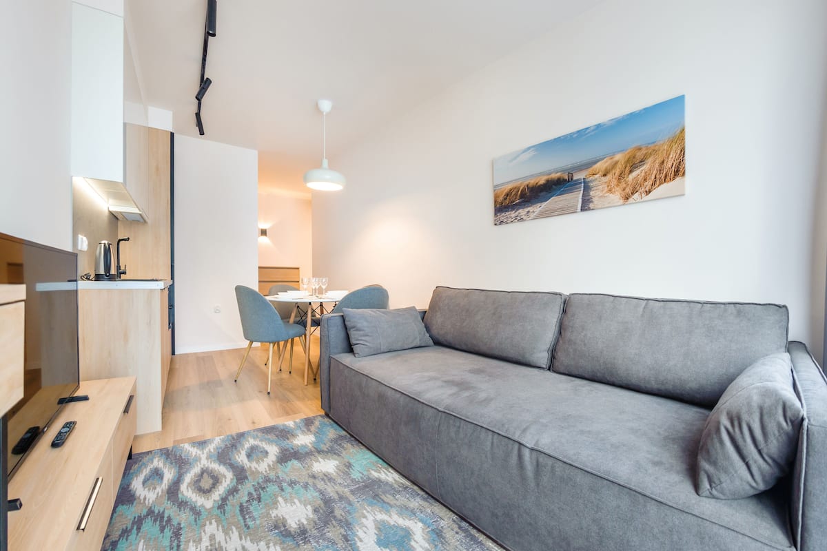 A modern living room with a grey sofa, a patterned rug, a dining area with a table and chairs, and a beach-themed painting on the wall.
