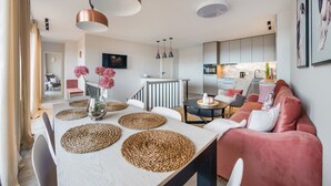 Una sala de estar moderna con comedor, que presenta una mesa de color claro, individuales tejidos, un sofá rosa e iluminación colgante.