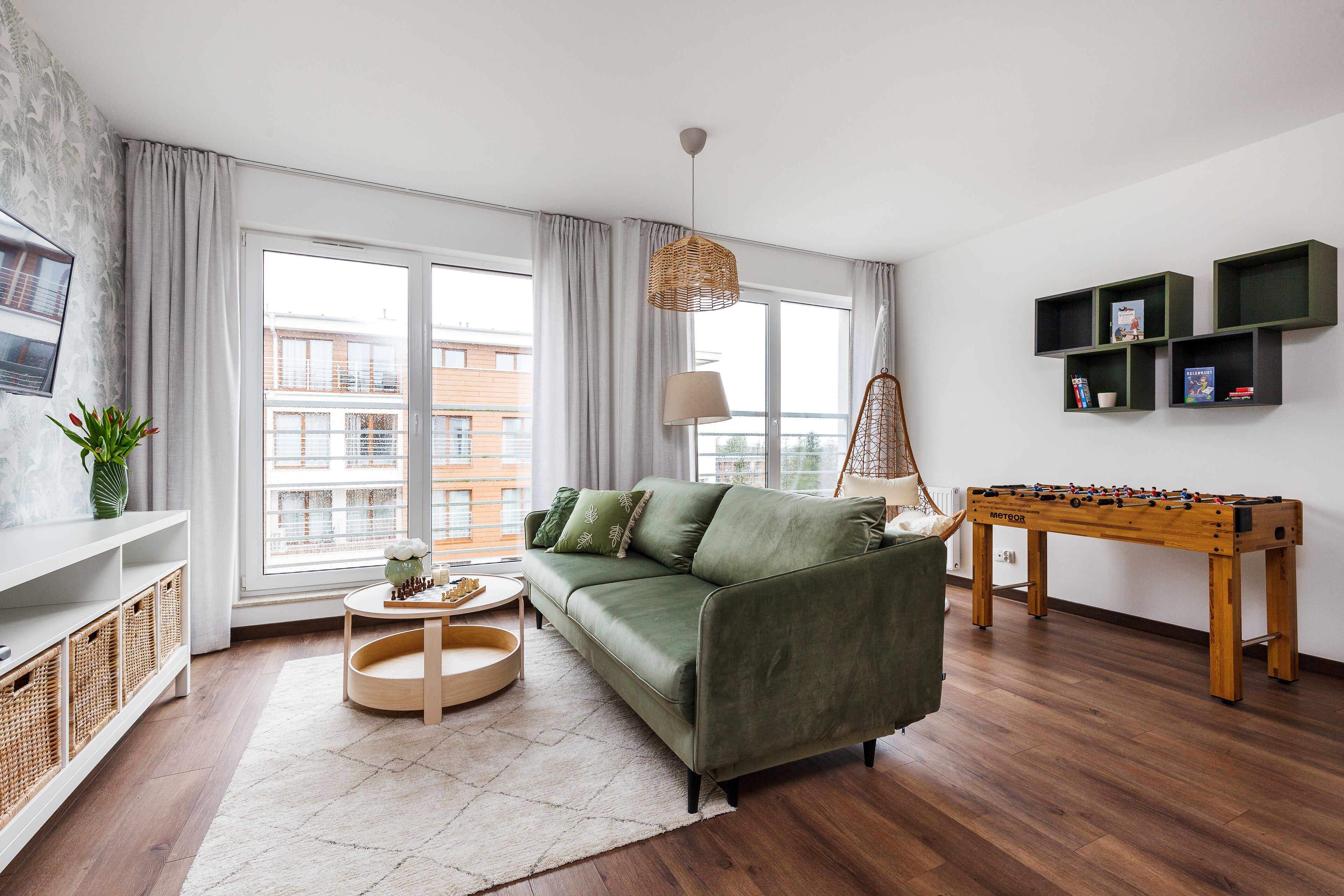 A modern living room with a green sofa, a foosball table, a TV, and a large window with curtains.