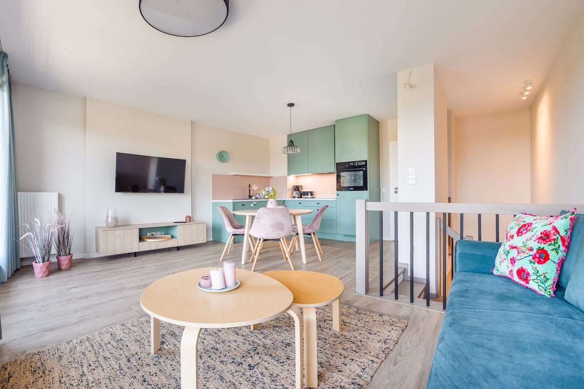 A modern living room with a blue sofa, a dining area with two chairs, a kitchen with teal cabinets, and a TV mounted on the wall.