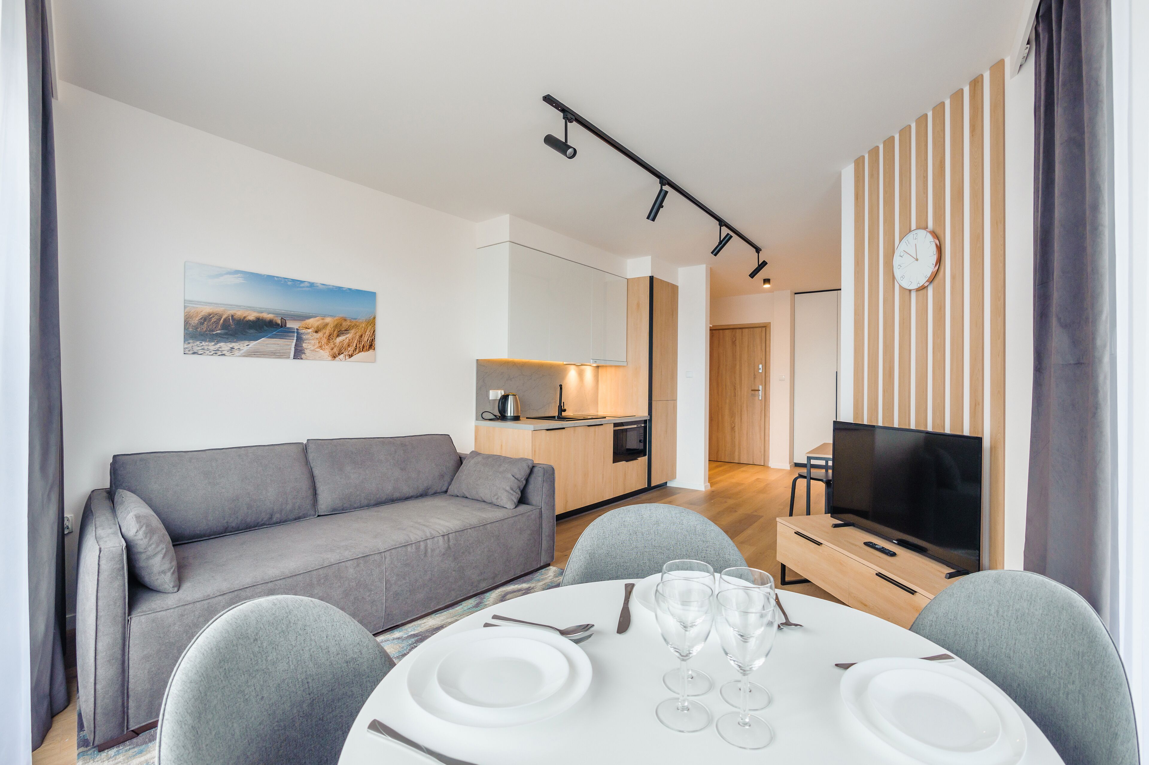 A modern living room with a grey sofa, a dining table set for two, and a kitchen in the background.