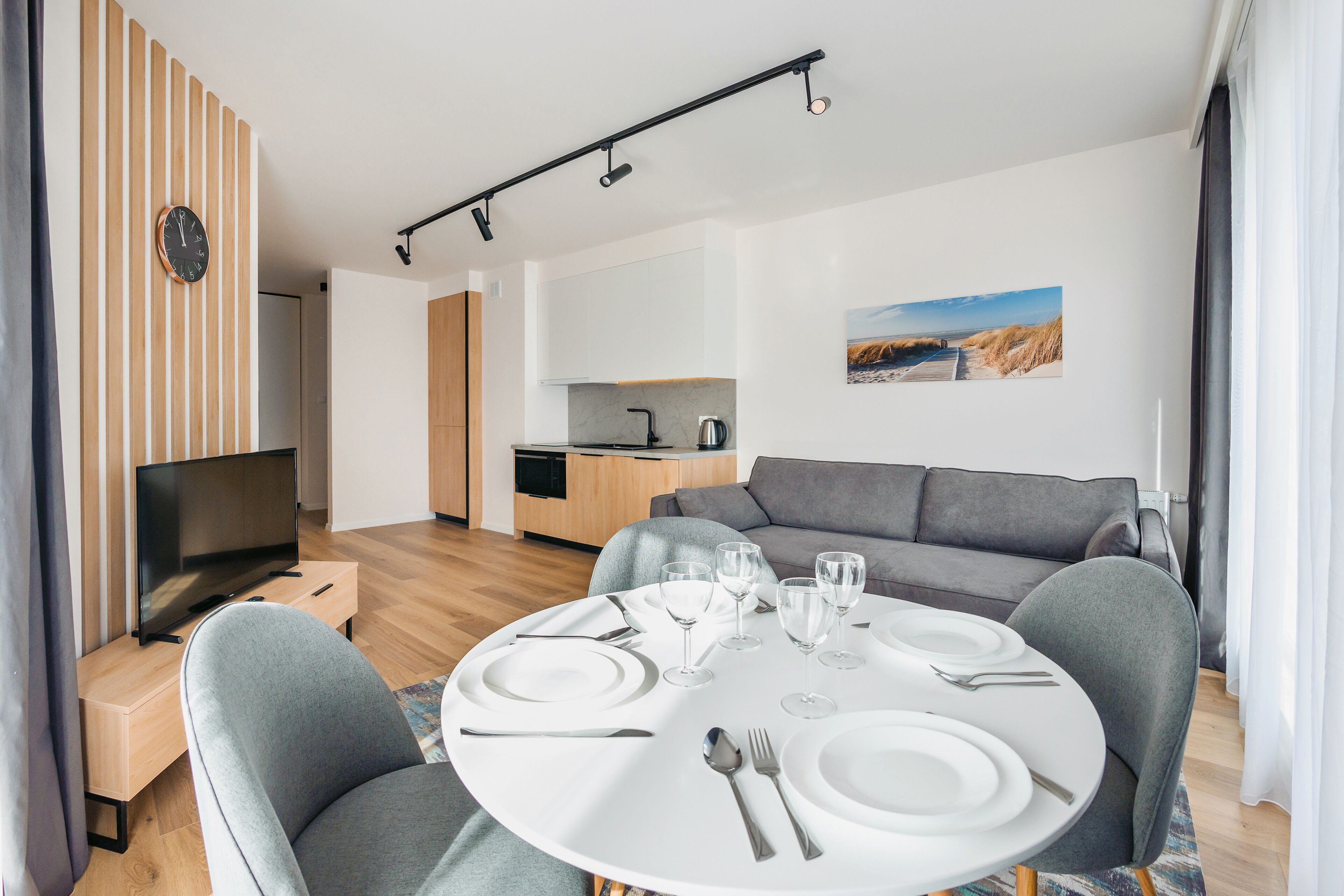A modern living room with a dining table set for four, a gray sofa, a flat-screen TV, and a kitchen in the background.