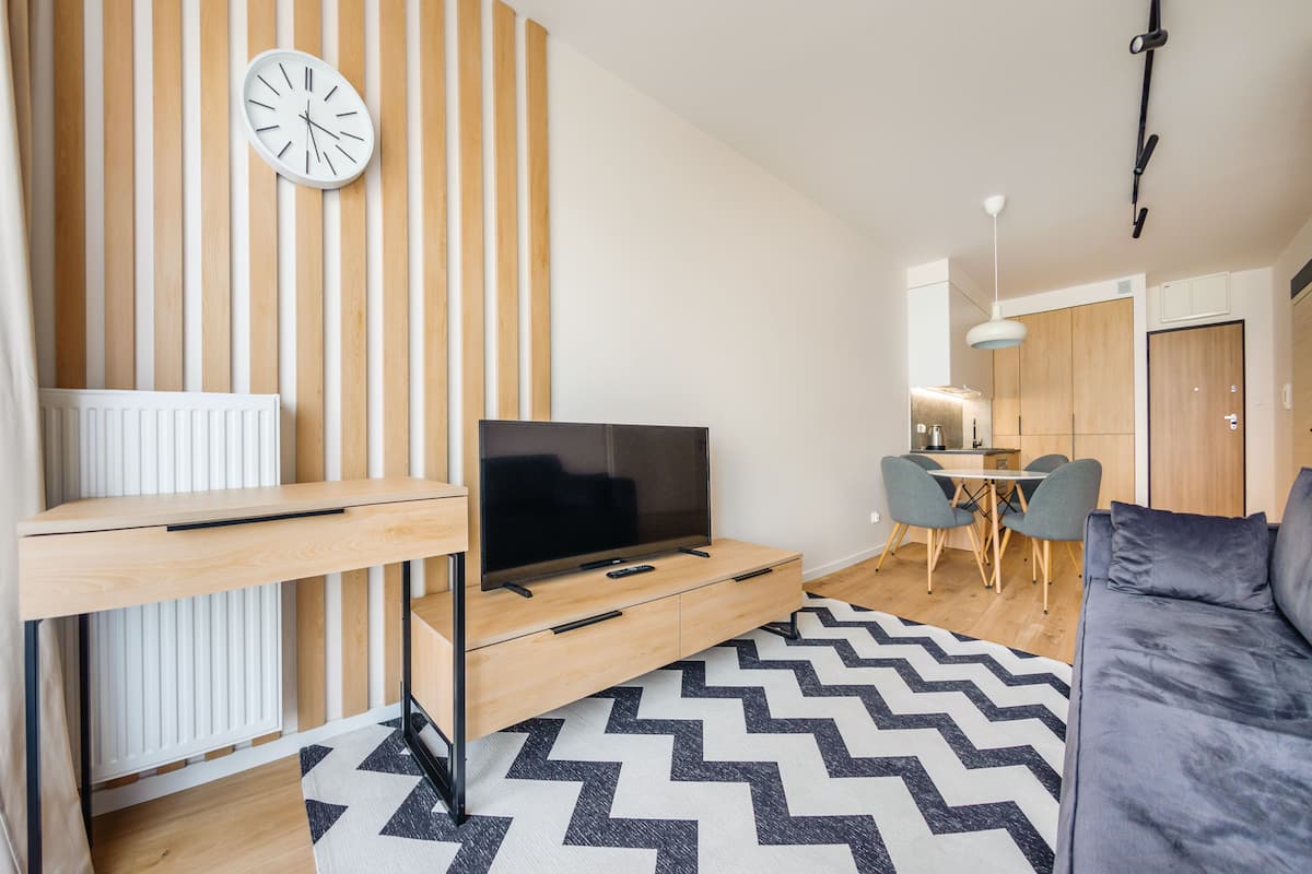 A modern living room with a TV on a wooden stand, a dining area with a table and chairs, and a kitchen in the background.
