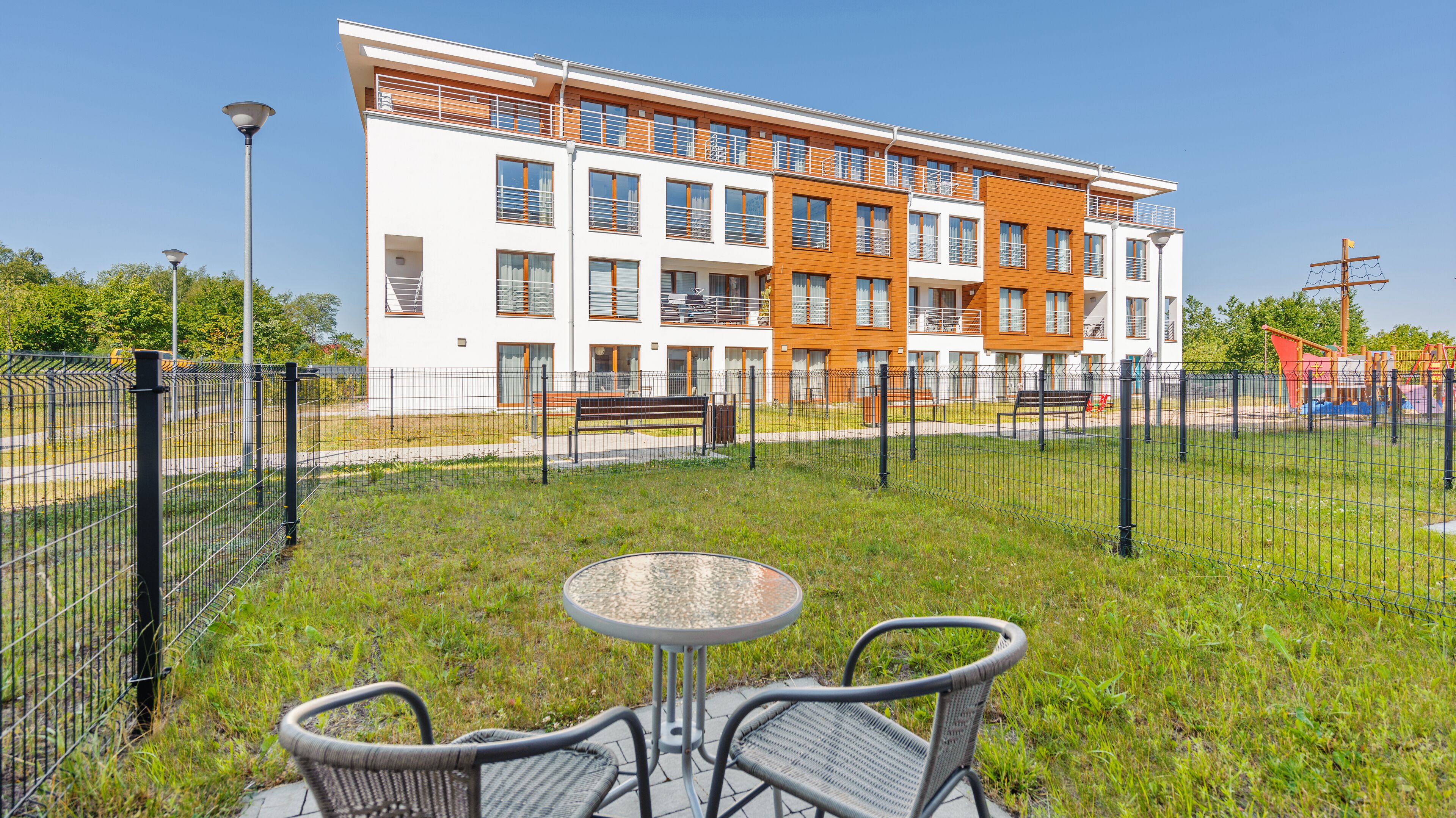 A modern building with multiple floors, outdoor seating area with metal chairs and a table, and a fenced playground in the background.