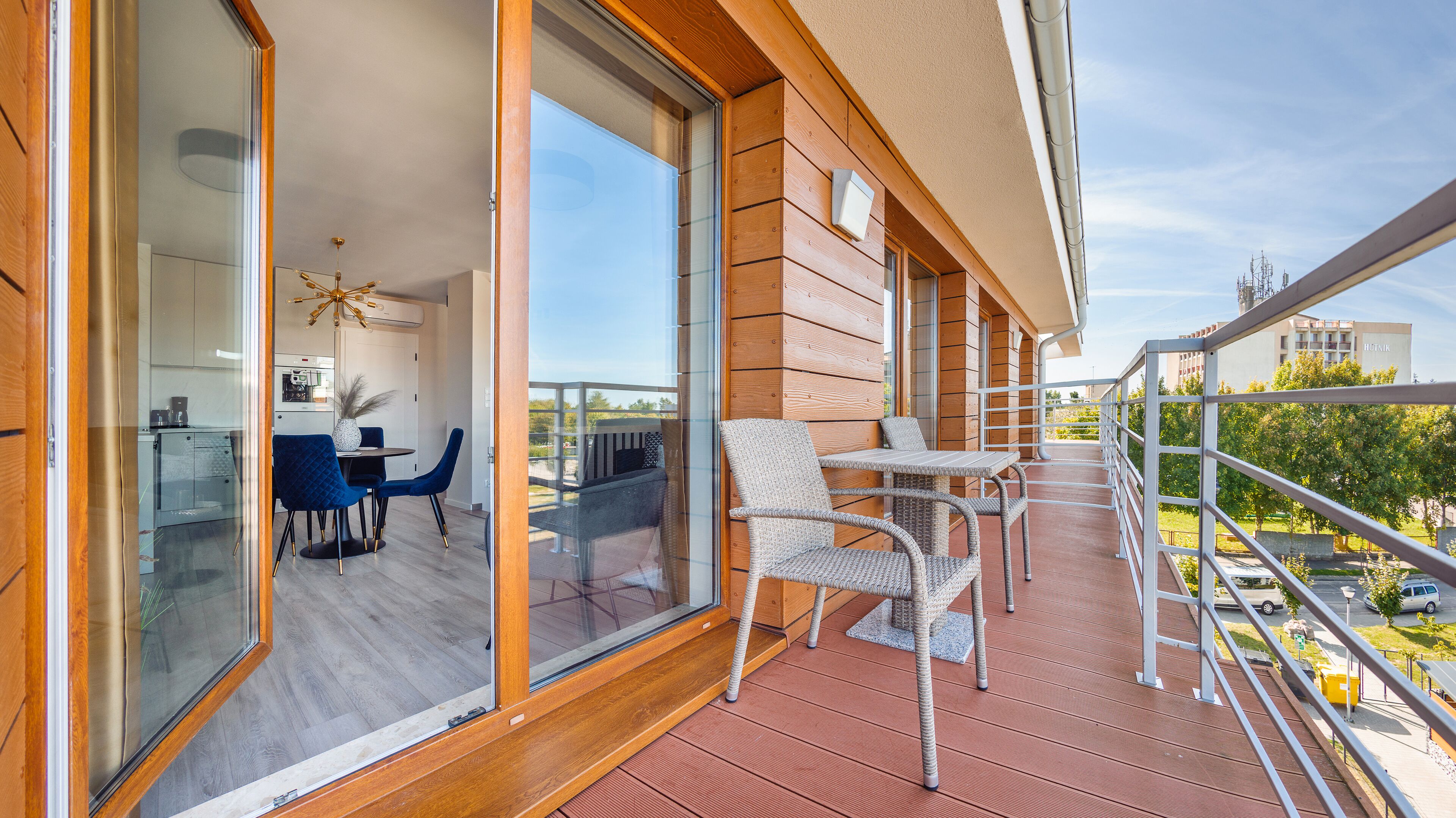 A balcony with wicker chairs, a glass door leading to a dining area with a table and chairs, and a view of the cityscape.