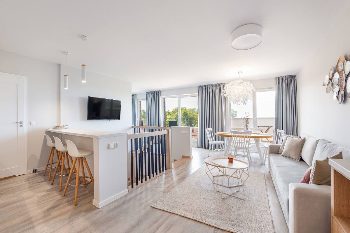 A modern apartment with a kitchen, dining area, and living room. It features a flat-screen TV, wooden bar stools, a dining table with chairs, and a sofa.