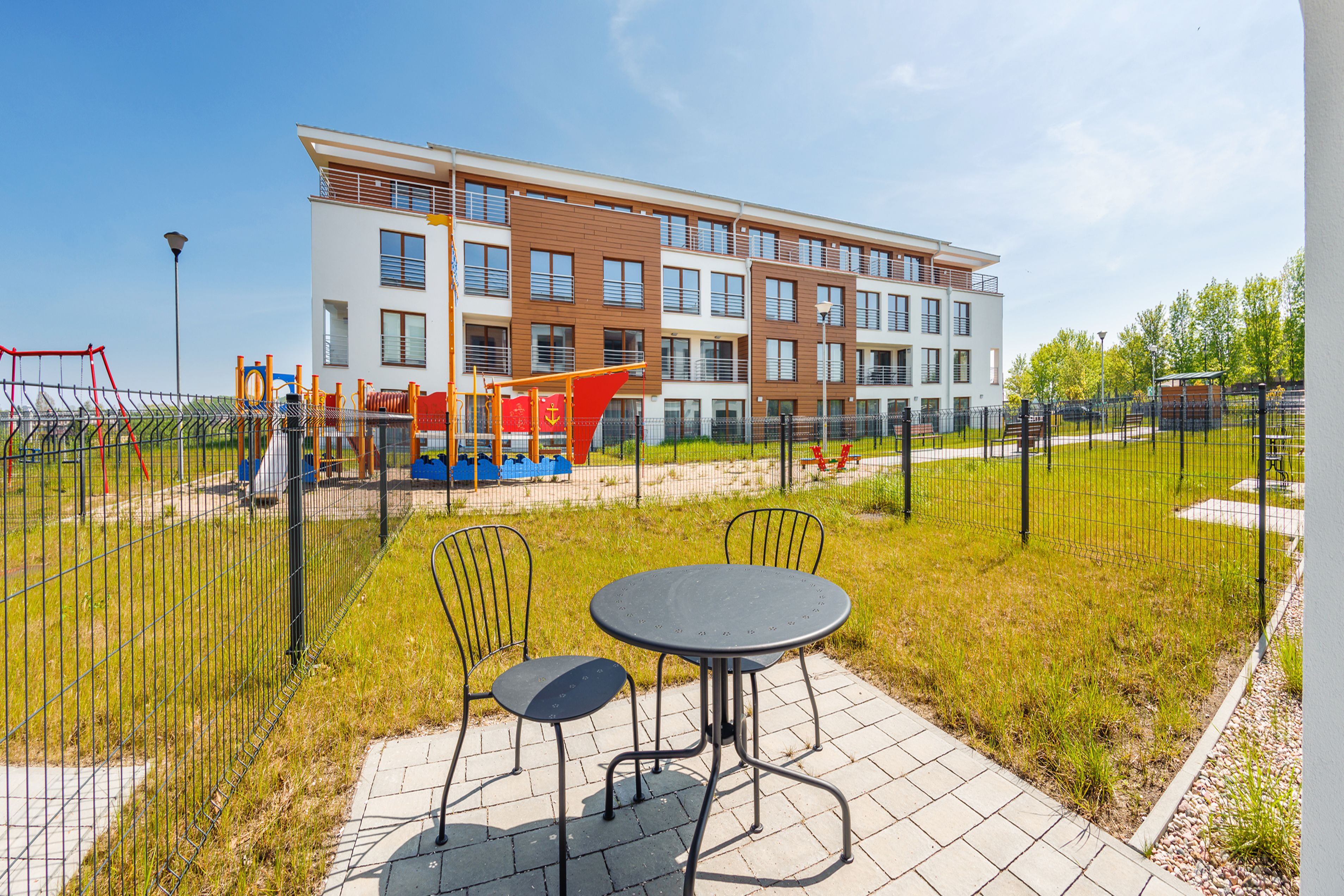 A playground area with a fenced play structure, a table with chairs, and a building in the background.