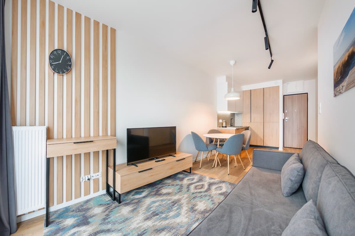 A modern living room with a flat-screen TV, a sofa, a dining area with chairs, and a kitchen in the background.
