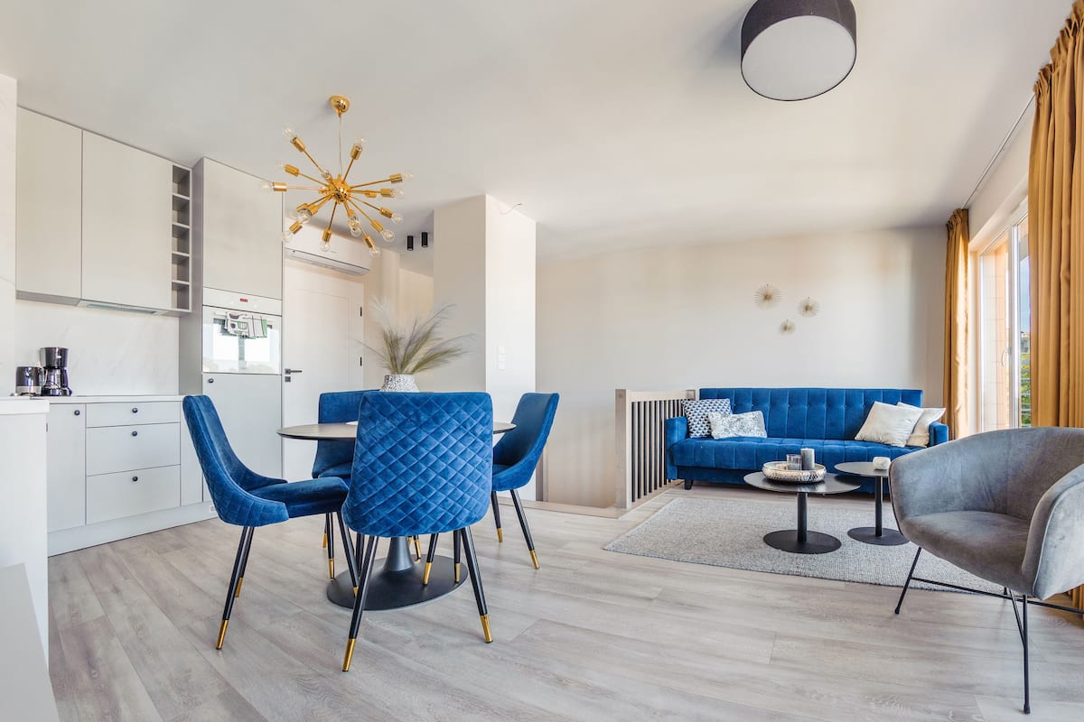 A modern living room with a blue sofa, a grey armchair, and a dining area with blue chairs.