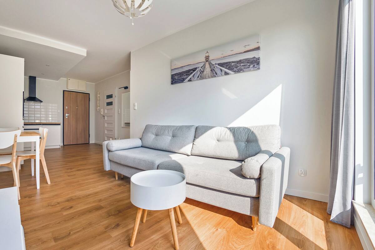 A modern living room with a grey sofa, wooden floor, and a dining area with white chairs.