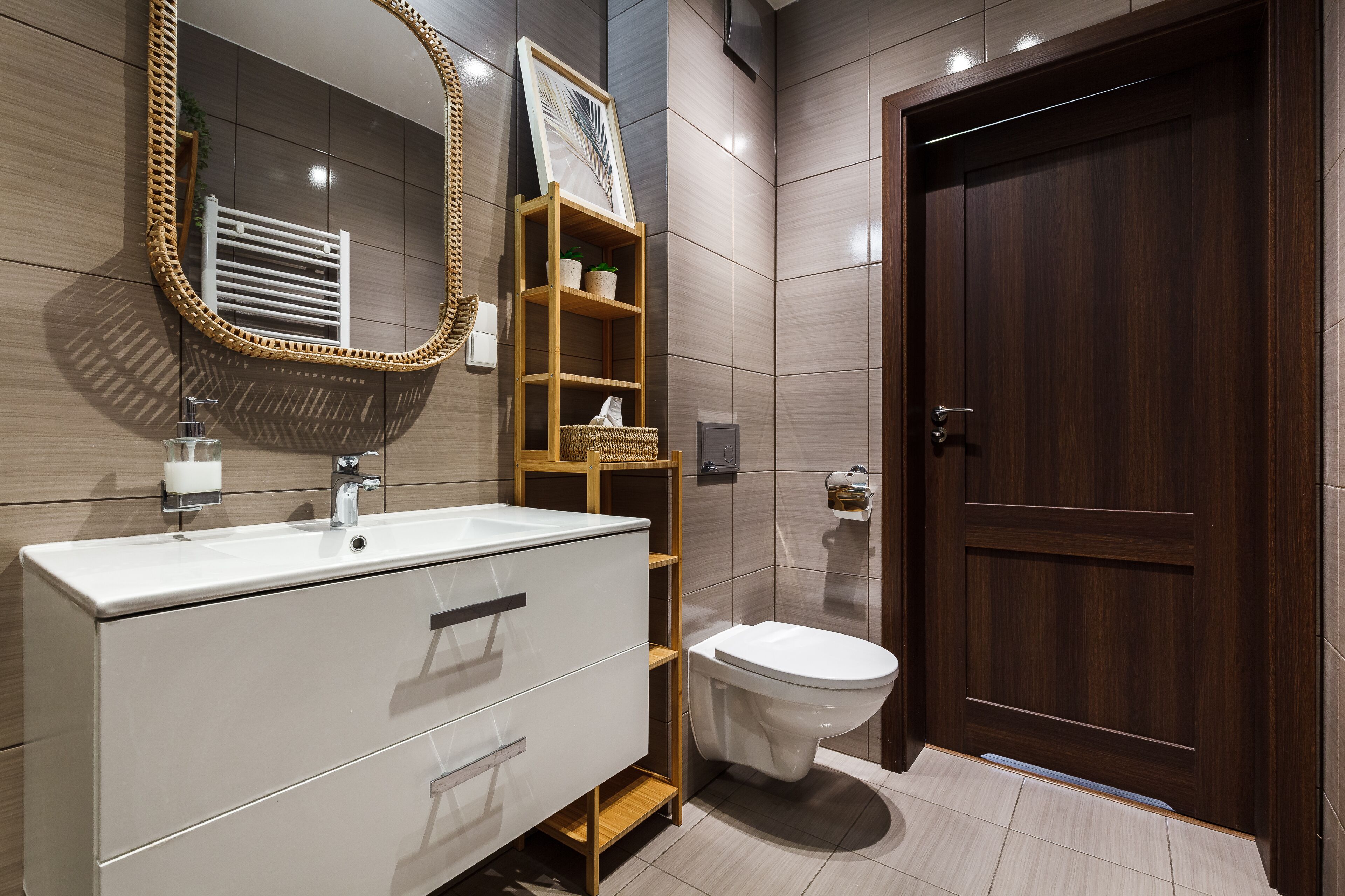 A modern bathroom with a white vanity, a wooden shelf with plants, a large mirror, and a dark wooden door.