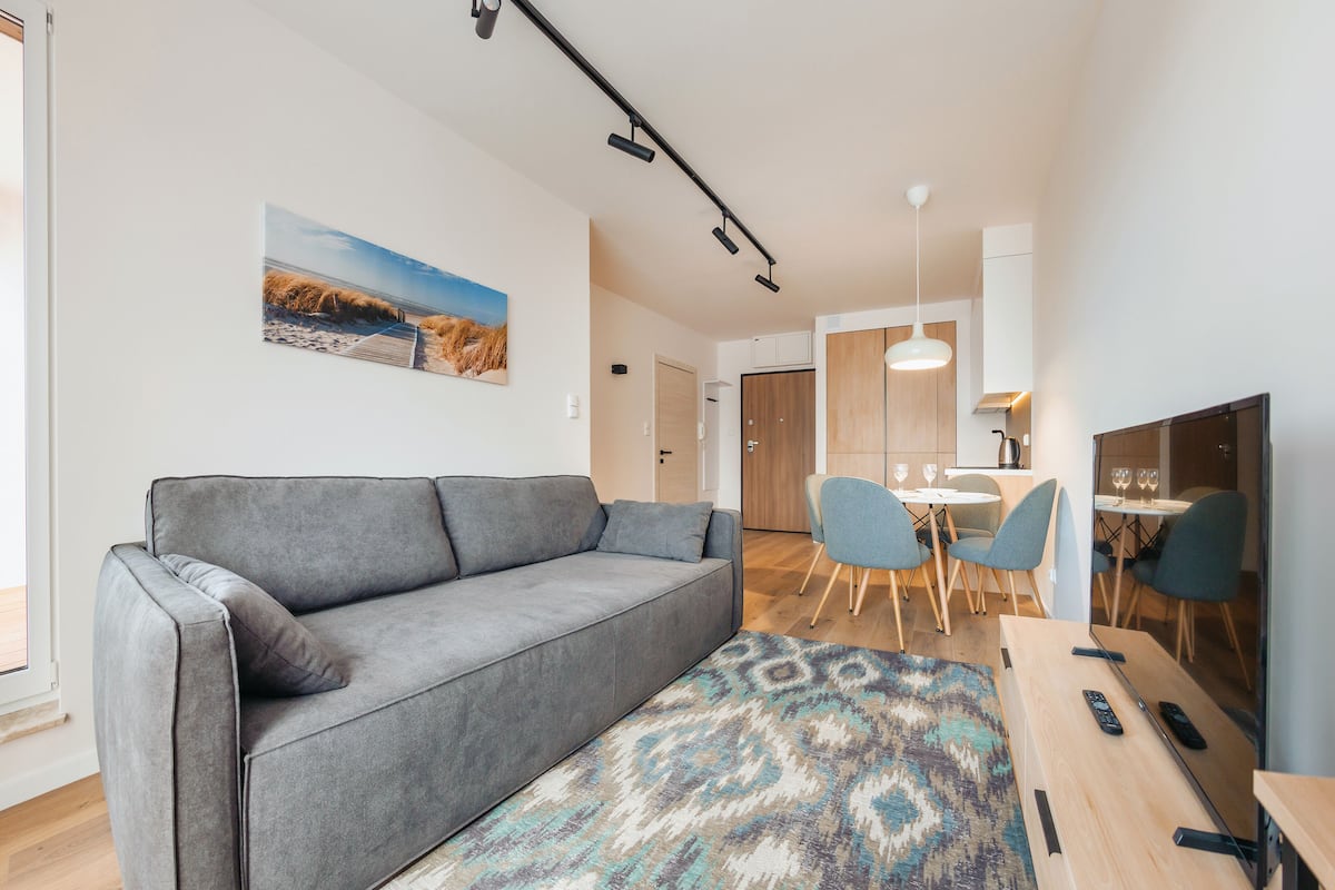 A modern living room with a grey sofa, a patterned rug, a wooden coffee table, and a dining area with chairs and a table.